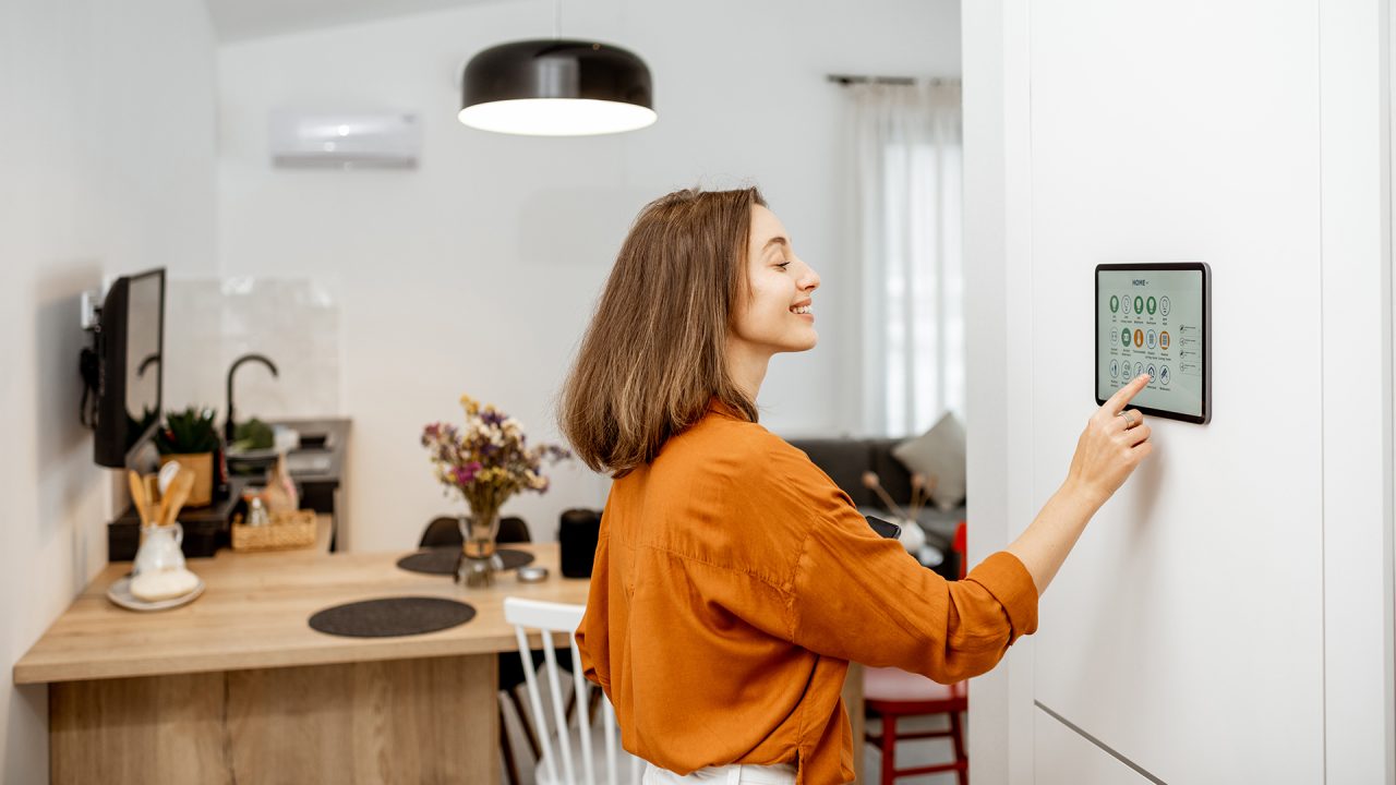 Young woman controlling home with a digital touch screen panel installed on the wall in the living room. Concept of a smart home and mobile application for managing smart devices at home